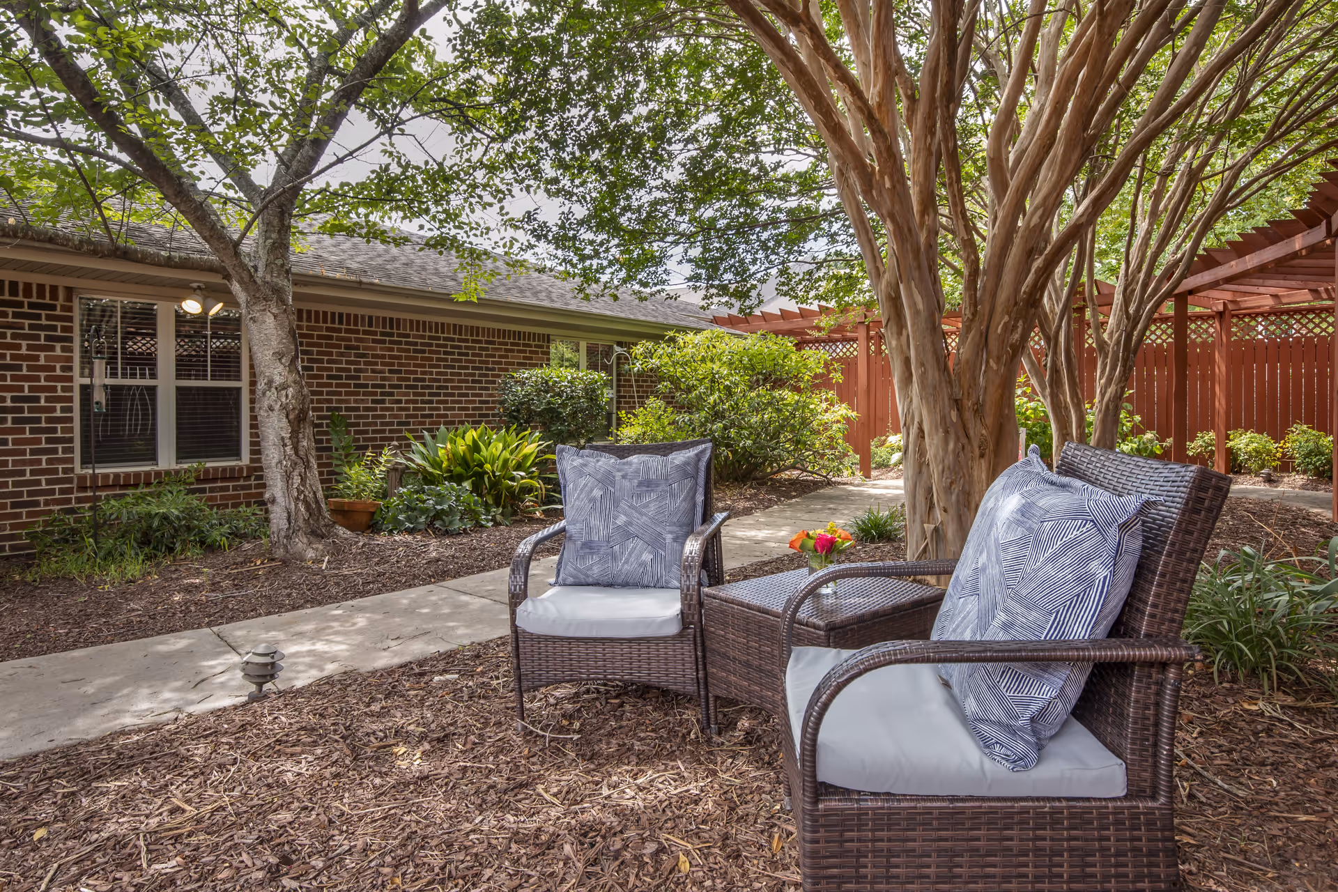 Outdoor seating area with two wicker chairs with cushions and a small table with a flower vase, surrounded by trees and plants, next to a brick building with windows and a red wooden fence in the background.