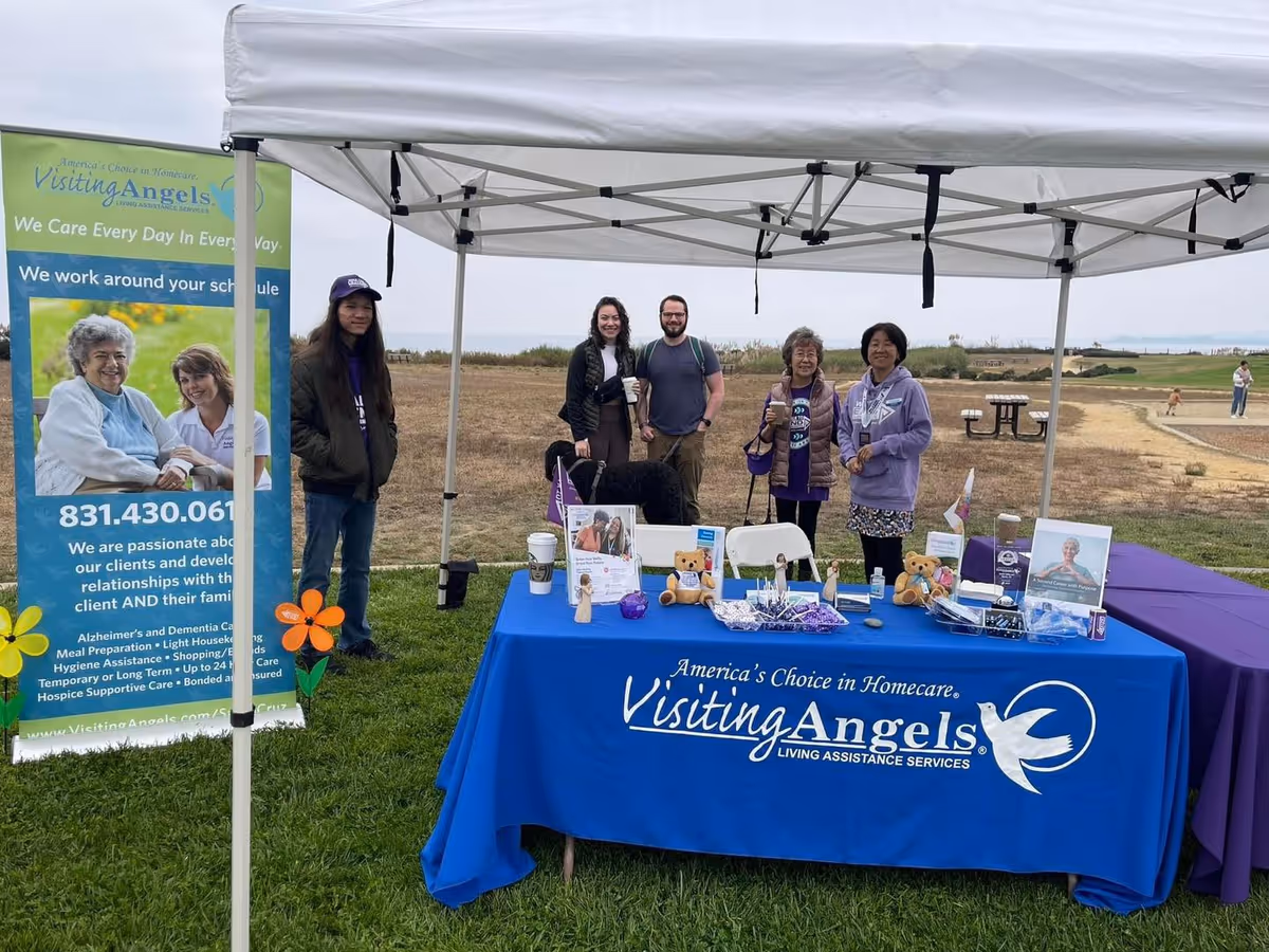 A group of people stands behind a blue Visiting Angels table under a white canopy outdoors.