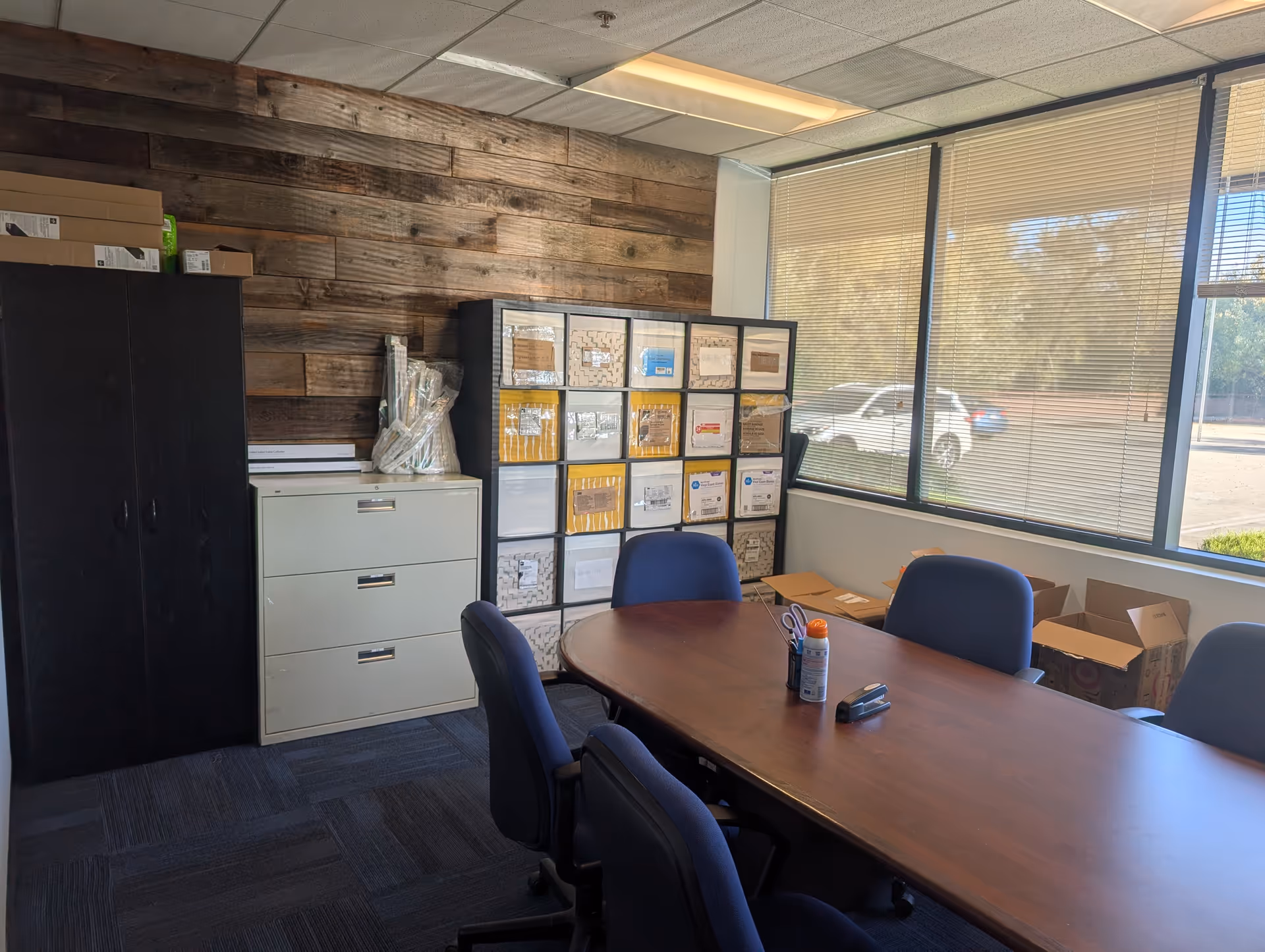 Office meeting room with table, chairs, filing cabinet, and storage shelves along a wood wall.