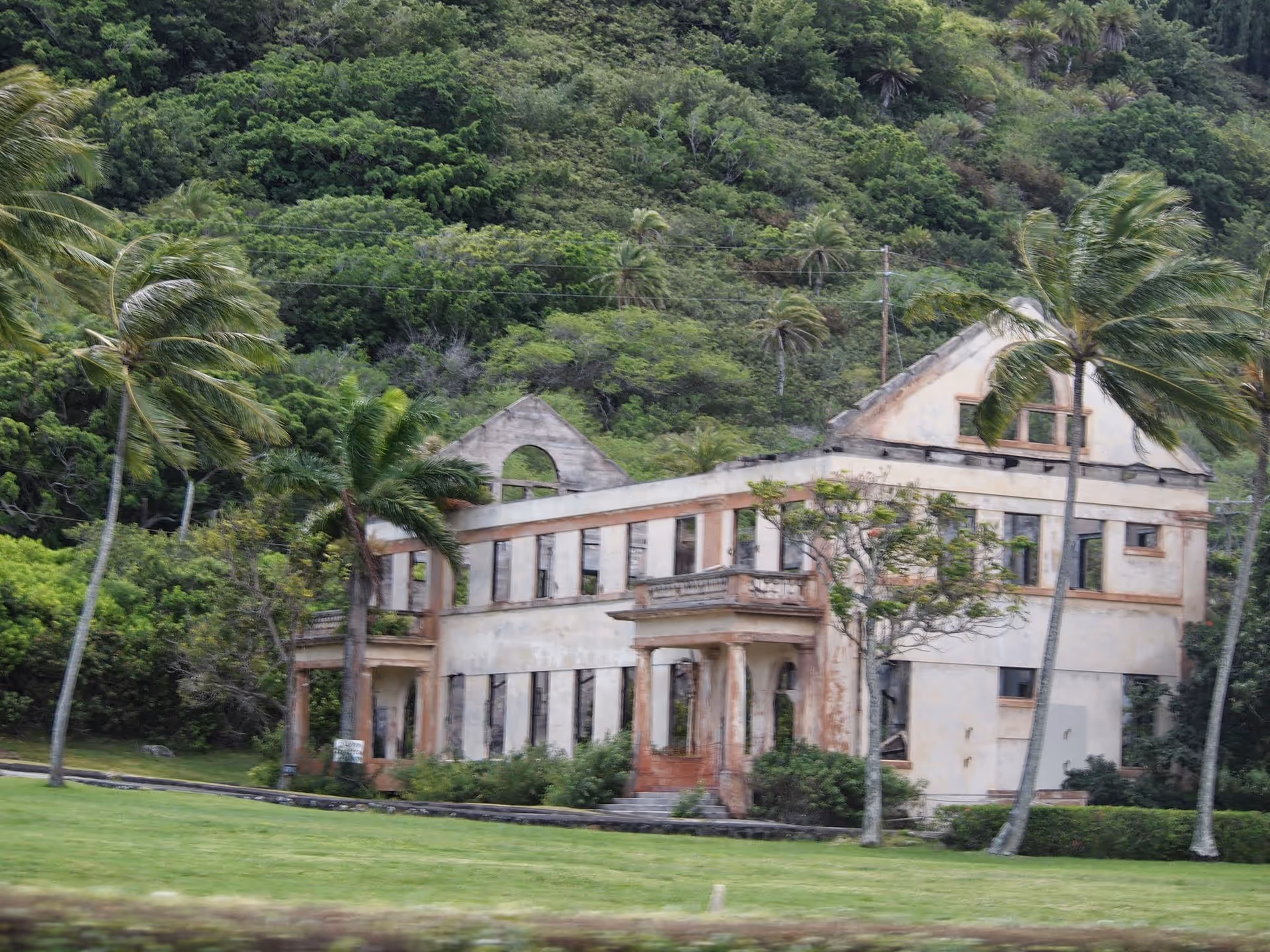 Dilapidated two-story building with arched windows surrounded by tropical trees and lawn.