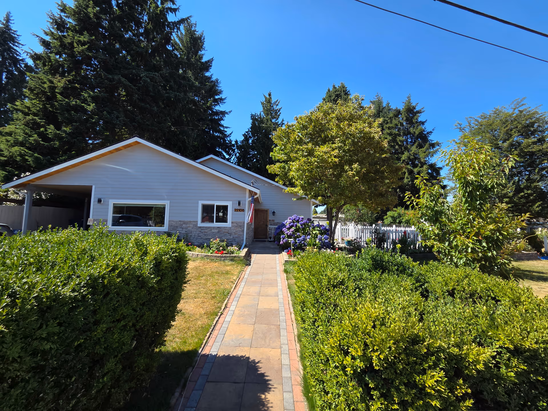 Single-story building with a paved path, trimmed hedges, and tall trees in a sunny yard.