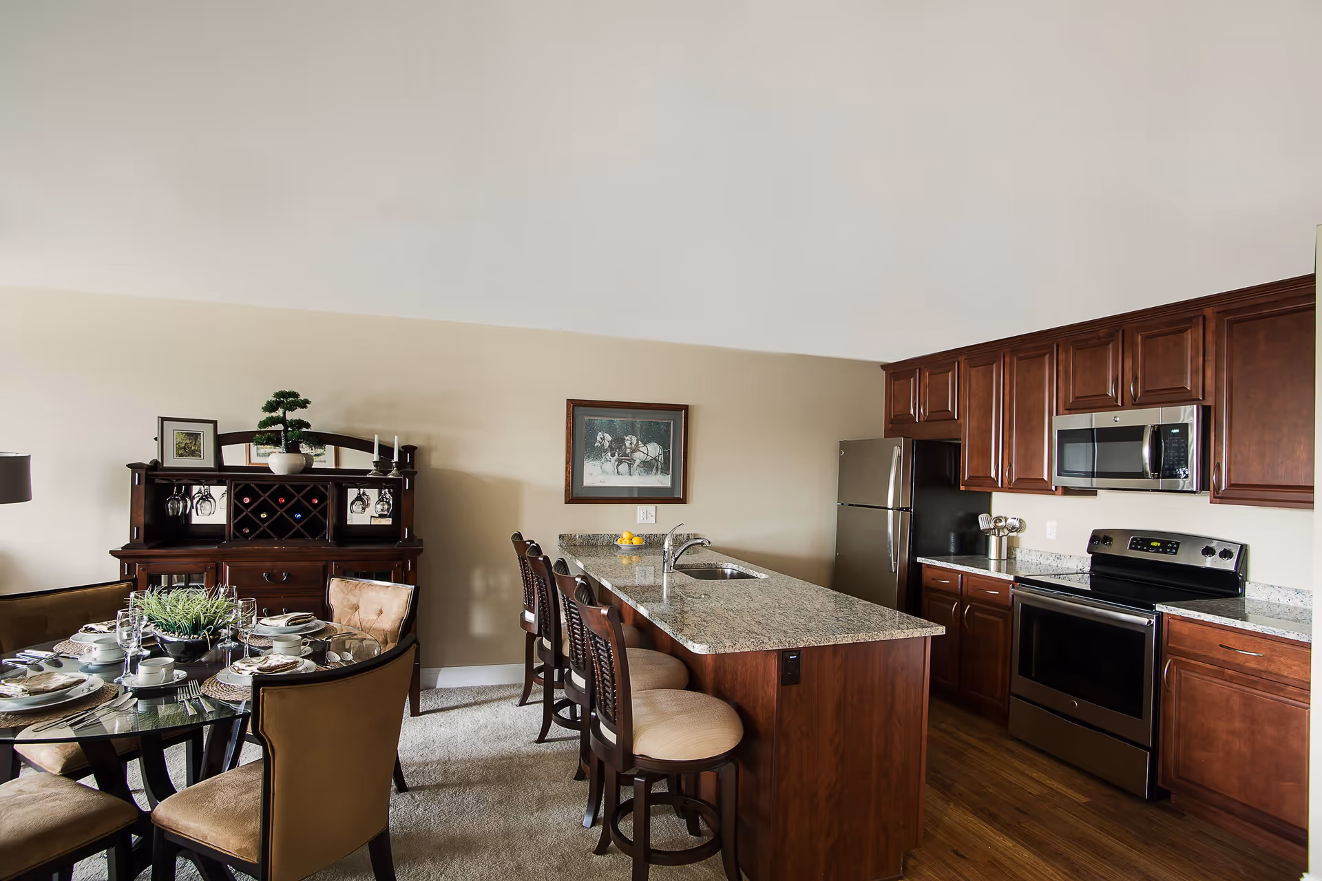 Interior view of a kitchen and dining area in Harrison Hill Apartments featuring a granite countertop island with four cushioned bar stools, stainless steel appliances including a refrigerator, microwave, and stove, wooden cabinets, a round glass dining table set with plates, cups, and cutlery, and a wooden hutch with wine storage and decorative items against a beige wall.