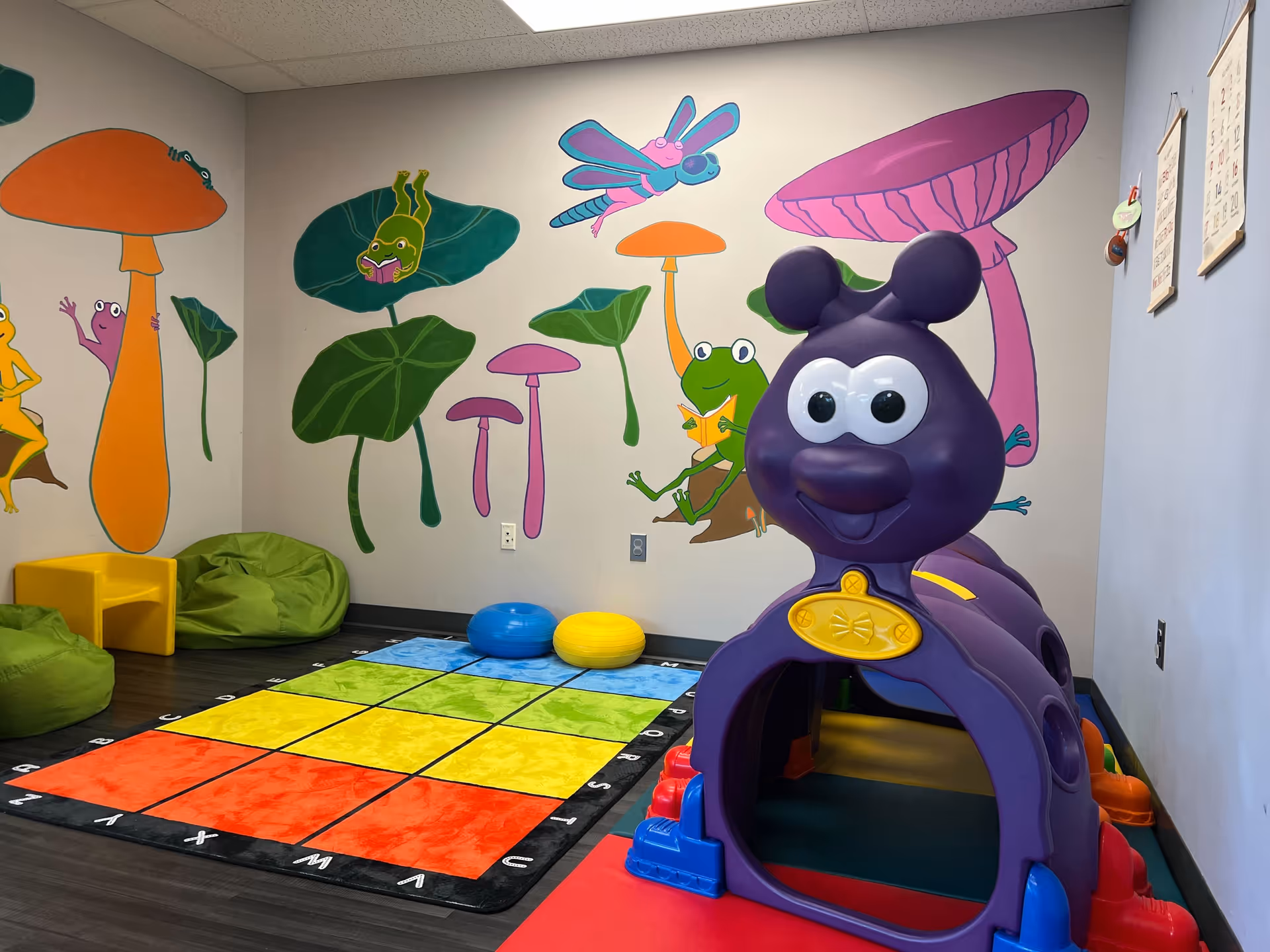 Colorful sensory play room with mural walls, soft seating, mats, and a large purple play structure.