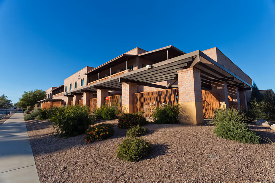 Exterior view of a modern senior living facility building with brick walls, large covered patio areas, and landscaped desert plants under a clear blue sky.