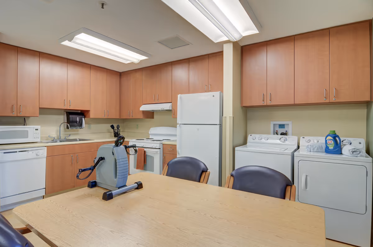 A communal kitchen in a senior living facility with wooden cabinets, appliances, and a table.