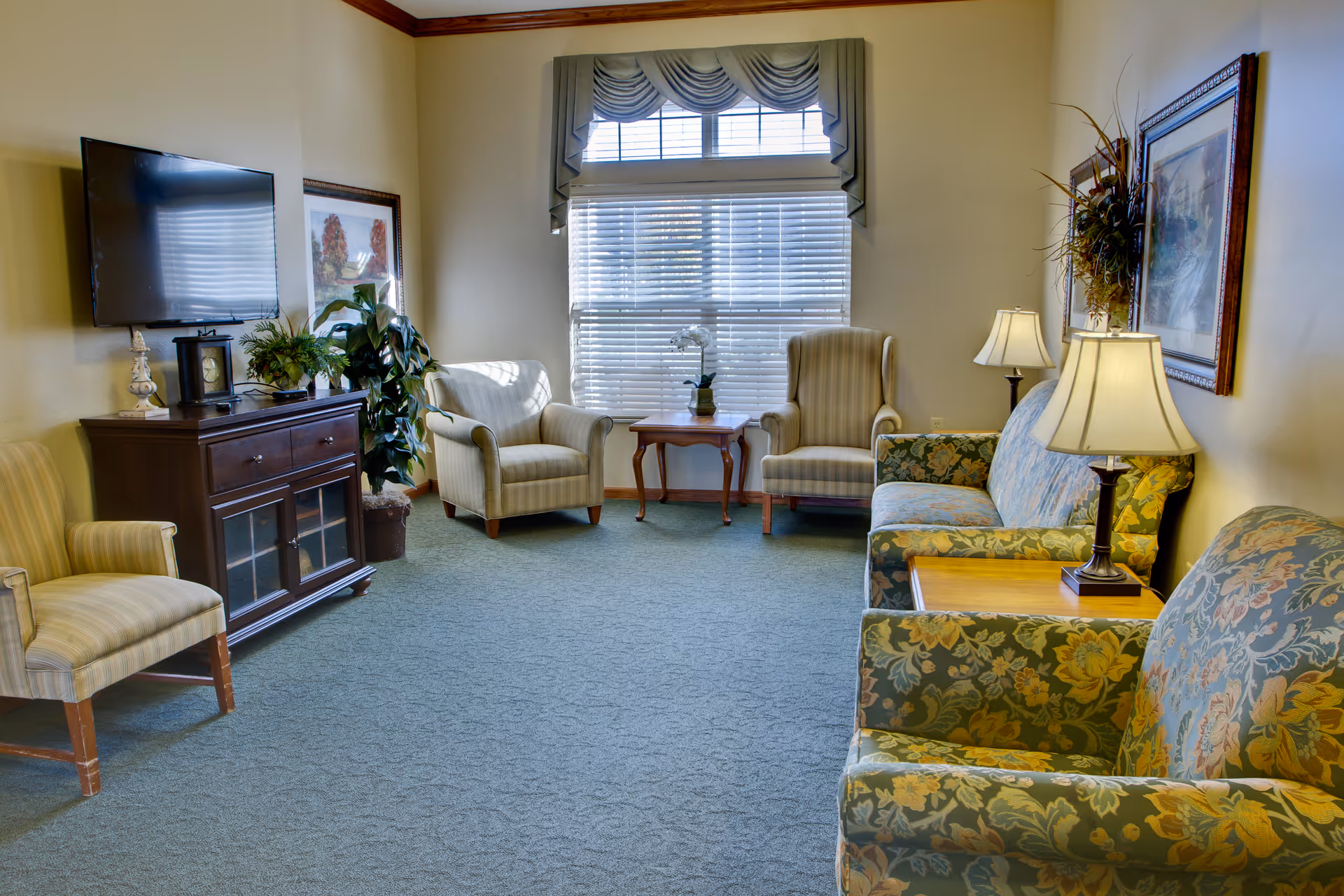 Cozy senior living lounge with upholstered chairs, a sofa, TV, plants, and a window.