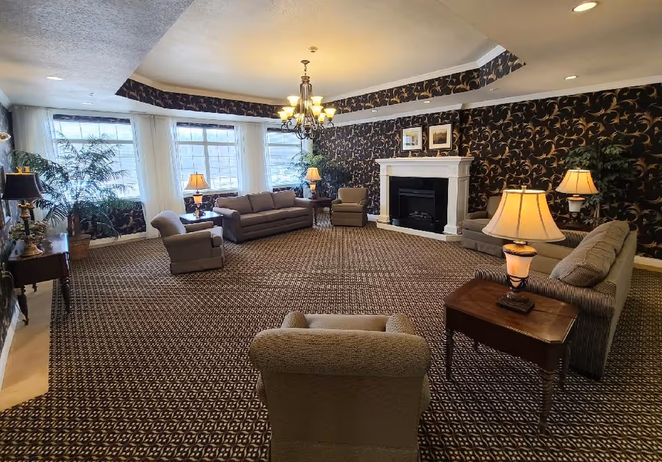 Spacious assisted-living common room with multiple sofas and armchairs arranged around a central fireplace, patterned carpet and wallpaper, large windows and table lamps.