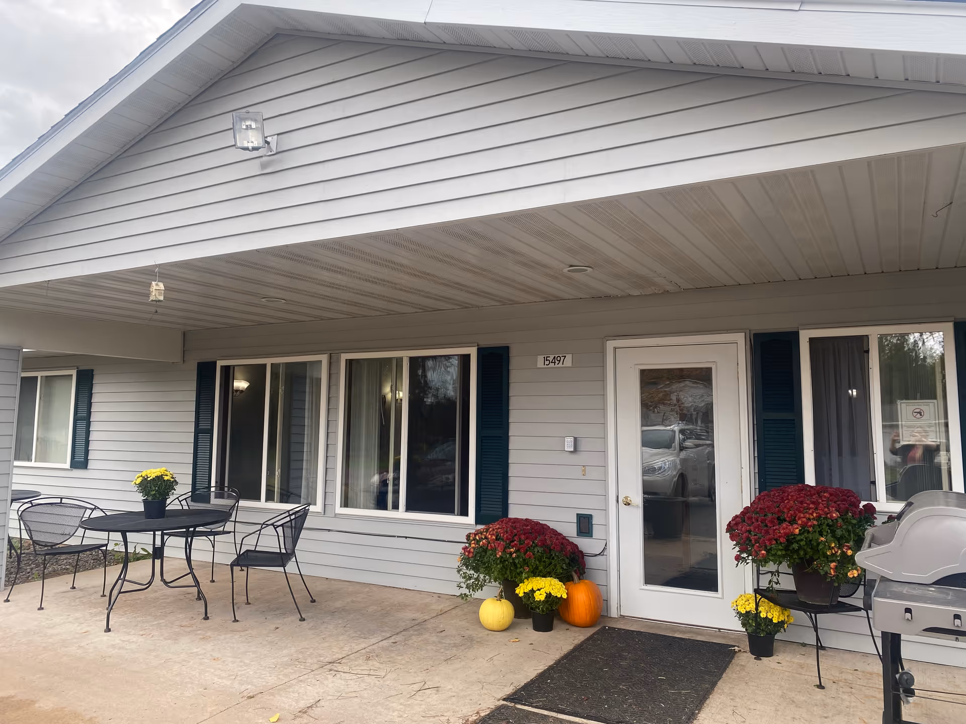 Exterior view of a single-story building with gray siding and a covered porch area. There is a white door with a glass panel, two windows with dark shutters, and a black metal table with four chairs. Several potted flowers and pumpkins are arranged near the door, and a grill is visible on the right side of the image. The building number 15497 is displayed above the door.
