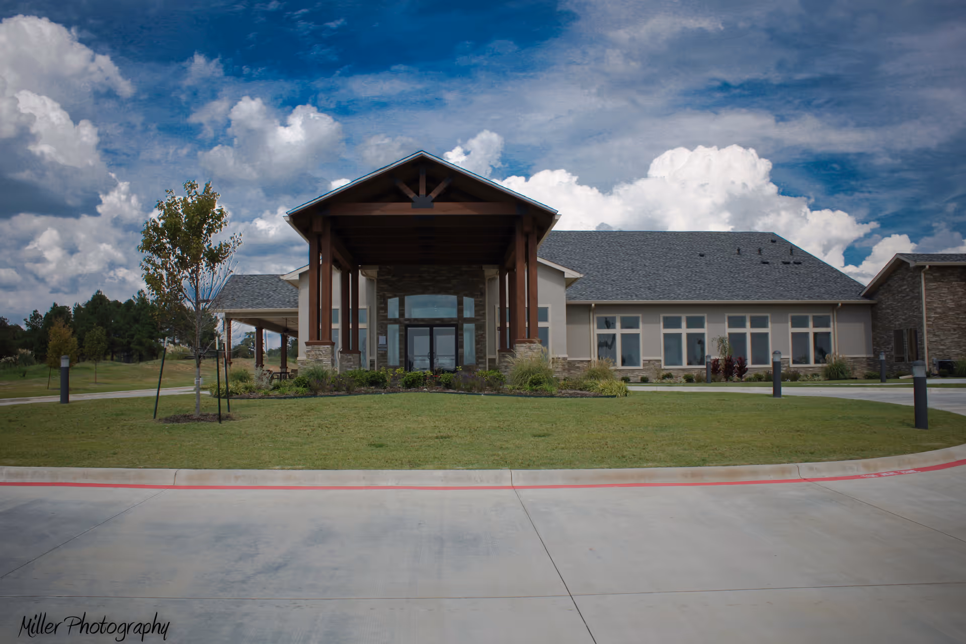 Front entrance of a single-story assisted living building with a covered porte-cochère, lawn, and cloudy sky.
