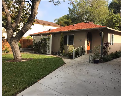 Single-story beige house with a red tile roof, front lawn, driveway, and a large tree in the yard.