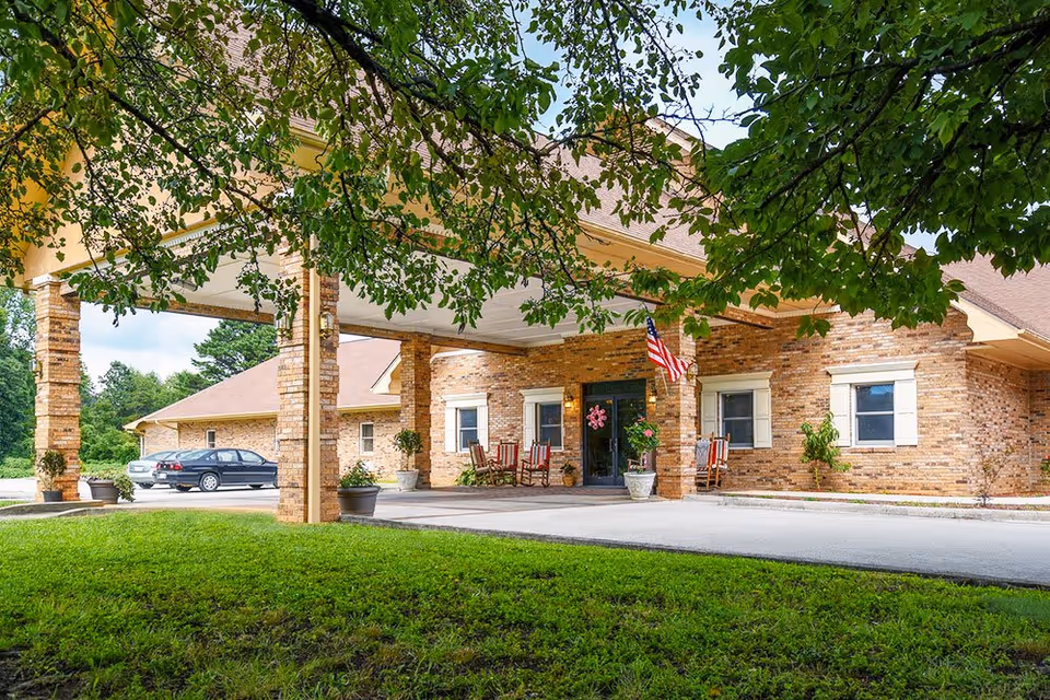 Covered brick entrance and porte-cochère of a senior living facility with rocking chairs, potted plants, an American flag, a parked car, and trees framing a grassy lawn.