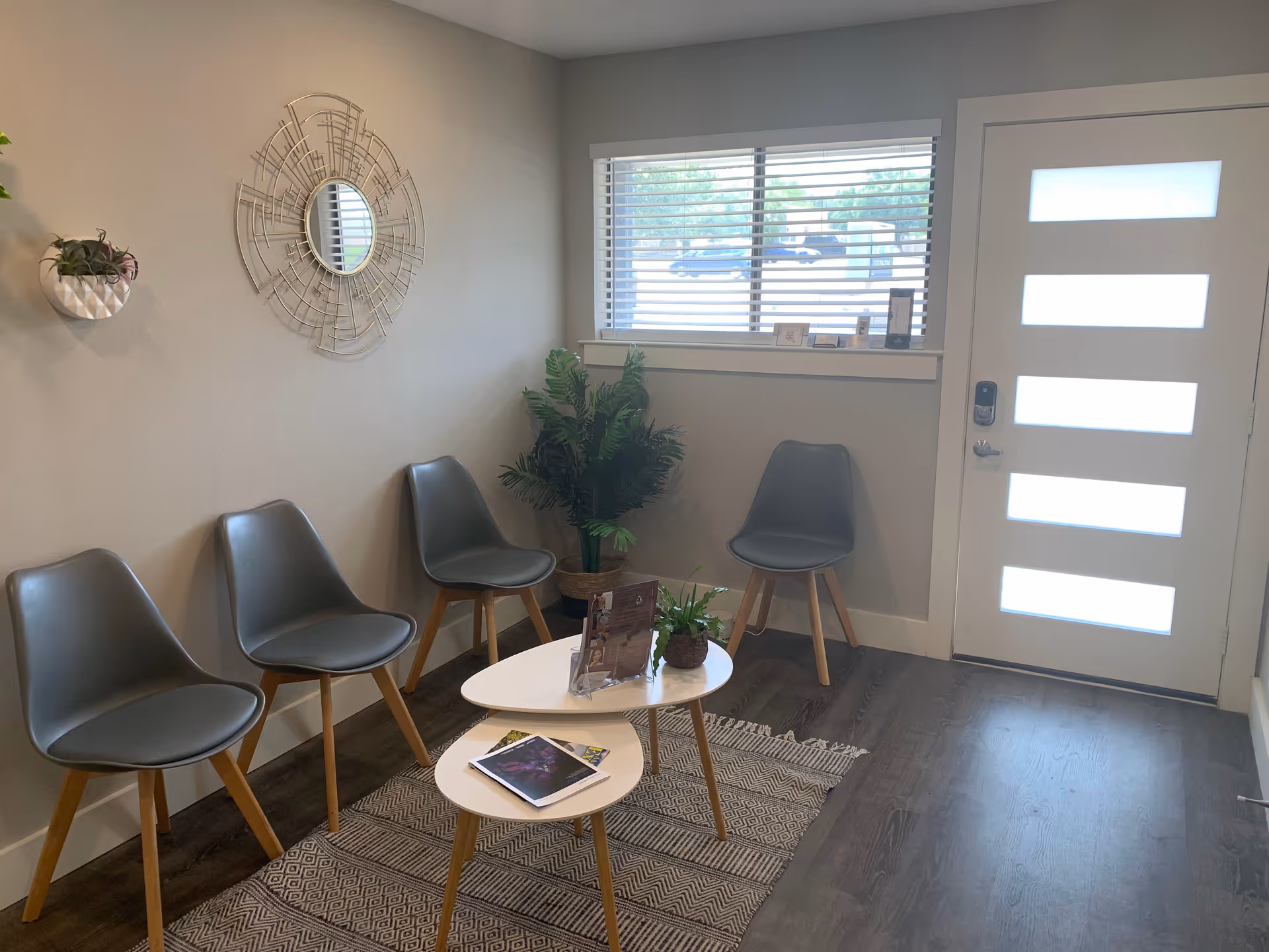 A small interior waiting area with gray chairs, white tables, plant decor, and a front door.