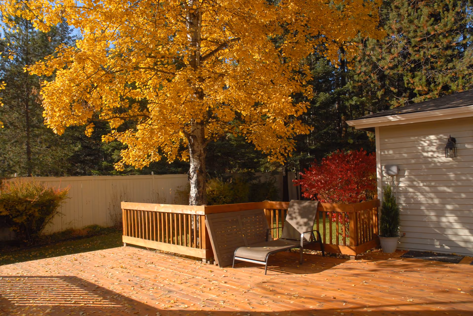 Outdoor wooden deck with a yellow autumn tree, seating, and a beige house exterior
