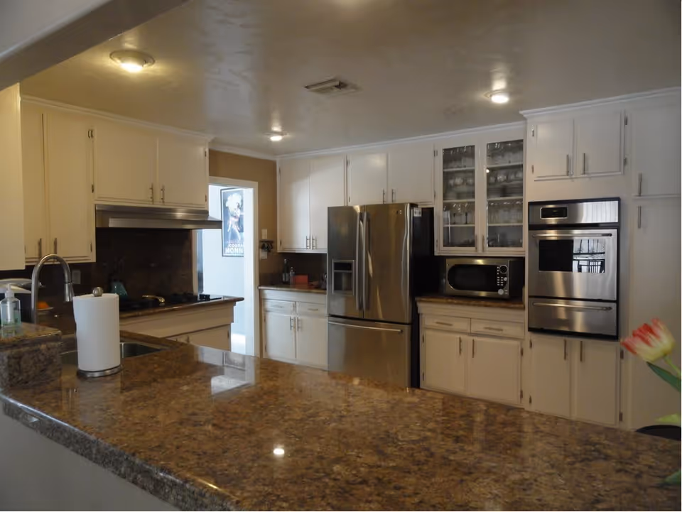 Interior view of a kitchen with white cabinets, a stainless steel refrigerator, built-in oven, microwave, and a granite countertop island with a paper towel holder and a sink. There is a doorway leading to another room with a poster on the wall.
