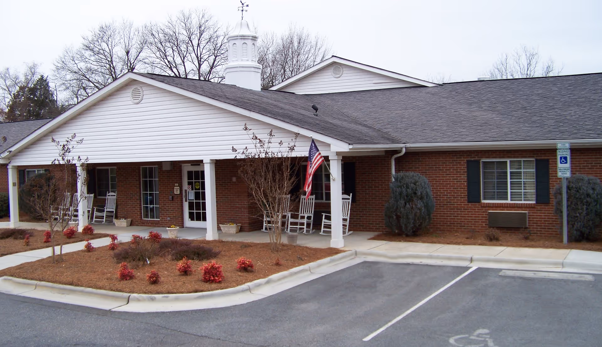 Exterior view of a single-story brick building with a white covered entrance supported by columns. There are rocking chairs on the porch, an American flag hanging near the entrance, small shrubs and plants in the landscaped area in front, and a parking lot with a handicapped parking space in the foreground. Leafless trees are visible in the background under an overcast sky.