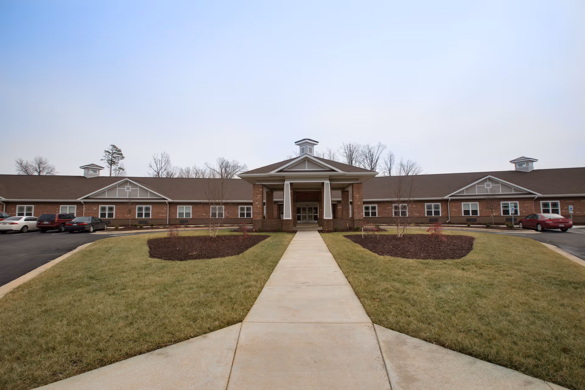 Front exterior view of Mebane Ridge Assisted Living facility showing a single-story brick building with a covered entrance, a paved walkway leading to the entrance, landscaped grass areas, and parked cars on either side.