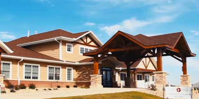 Front entrance of a senior living building with a large timber porte-cochere, beige siding, and landscaped grounds.