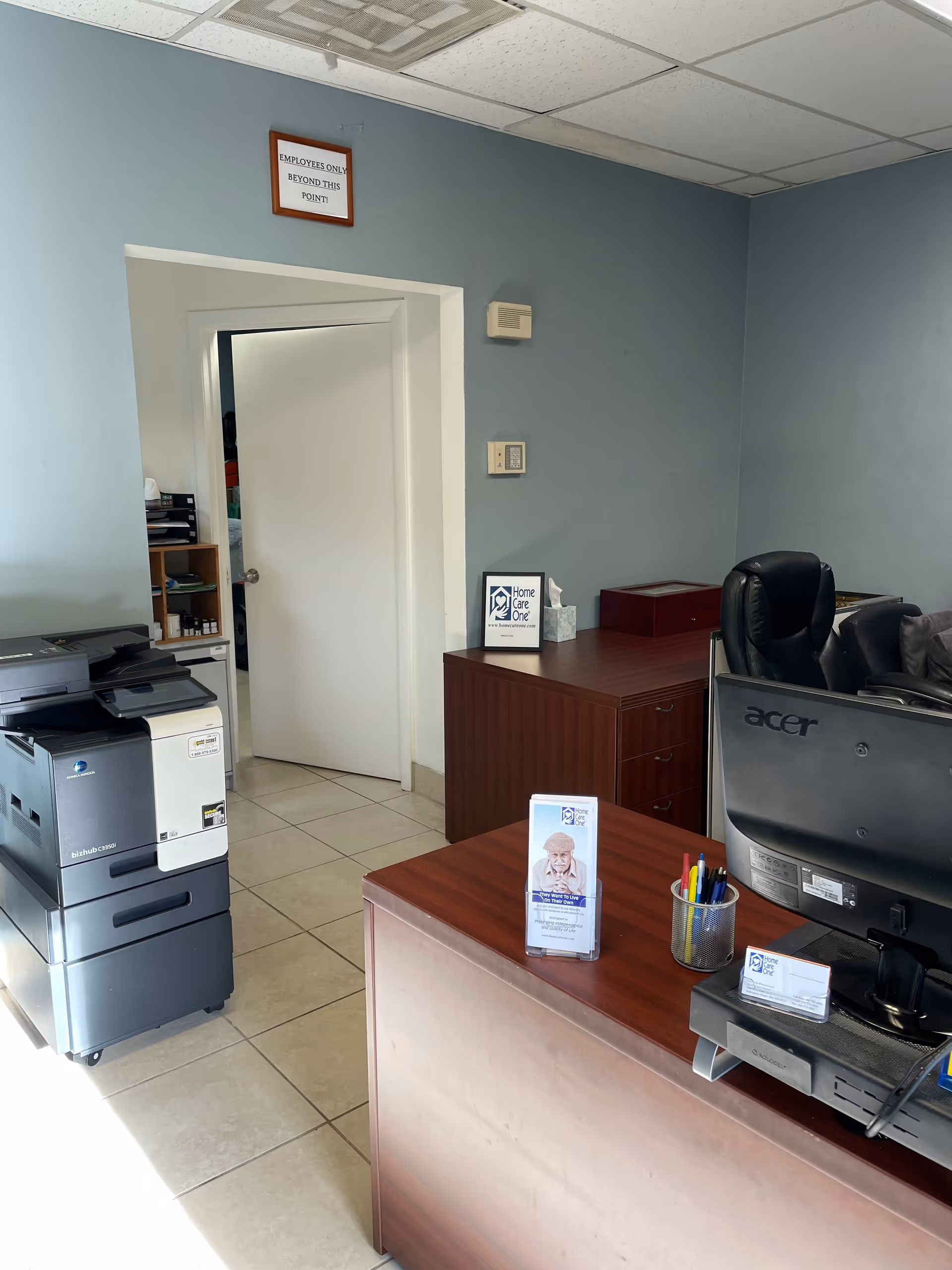 Office reception area with wooden desk, computer, printer, and doorway to another room.