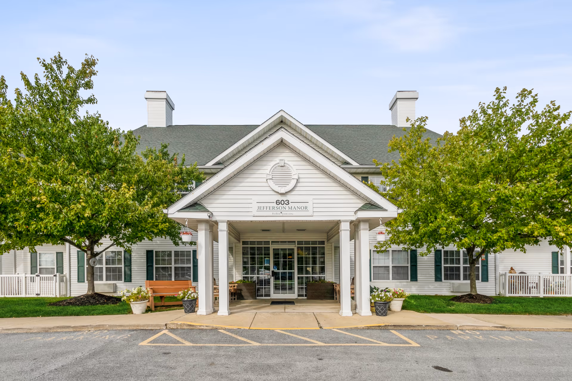 Front exterior view of Jefferson Manor, a white building with green shutters and a green roof. The entrance has a covered porch with white columns, and there are two trees with green leaves on either side of the entrance. There are benches and potted plants near the entrance.