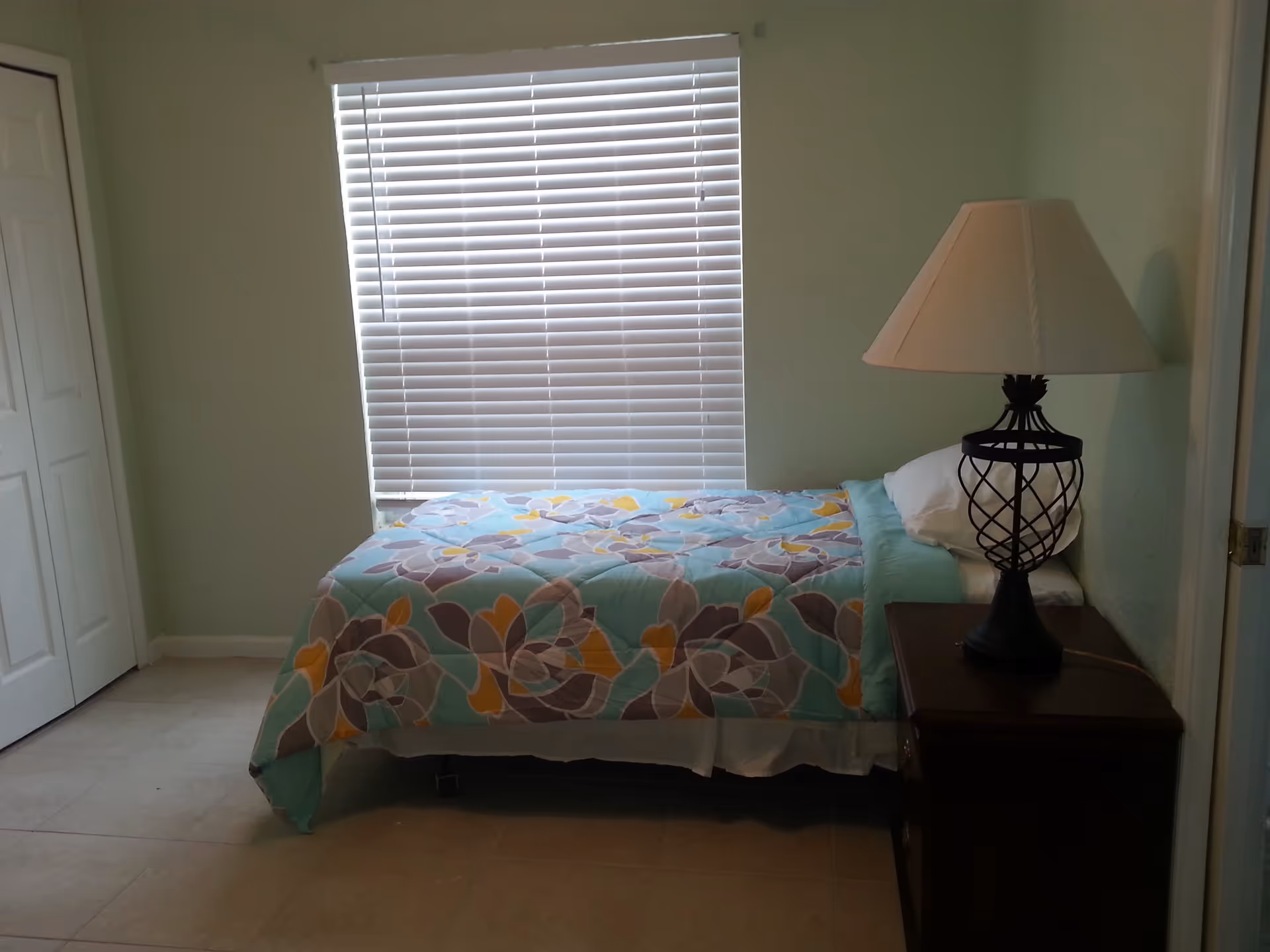 Small single bedroom with a floral-patterned bed under a window, a nightstand with a lamp, and a closet door.