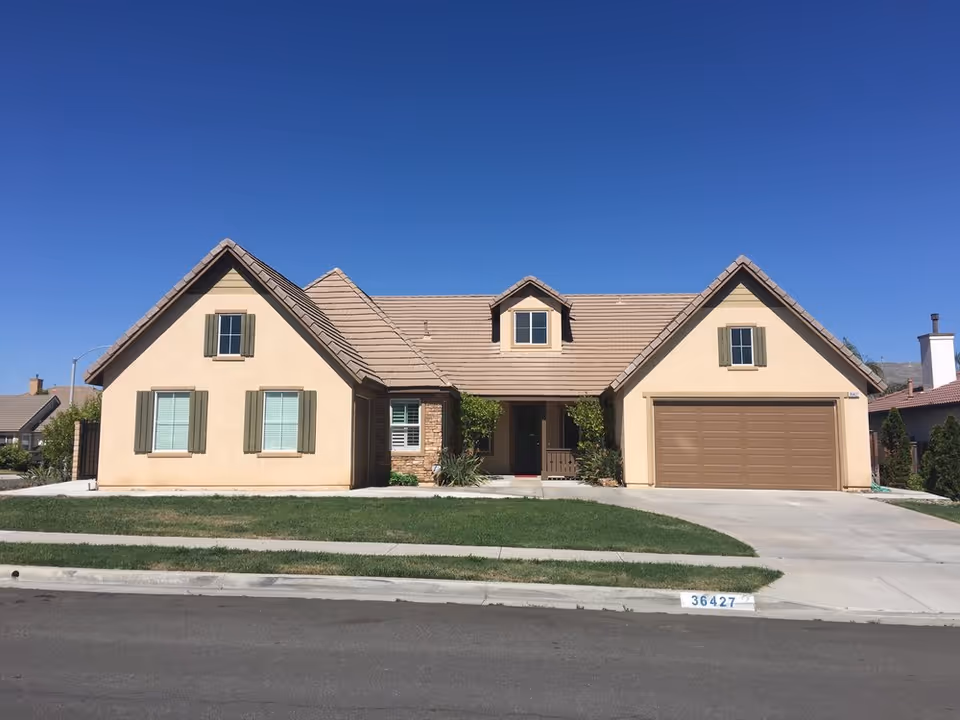 Single-story suburban house with beige stucco, an attached two-car garage, and a front lawn under a clear blue sky.