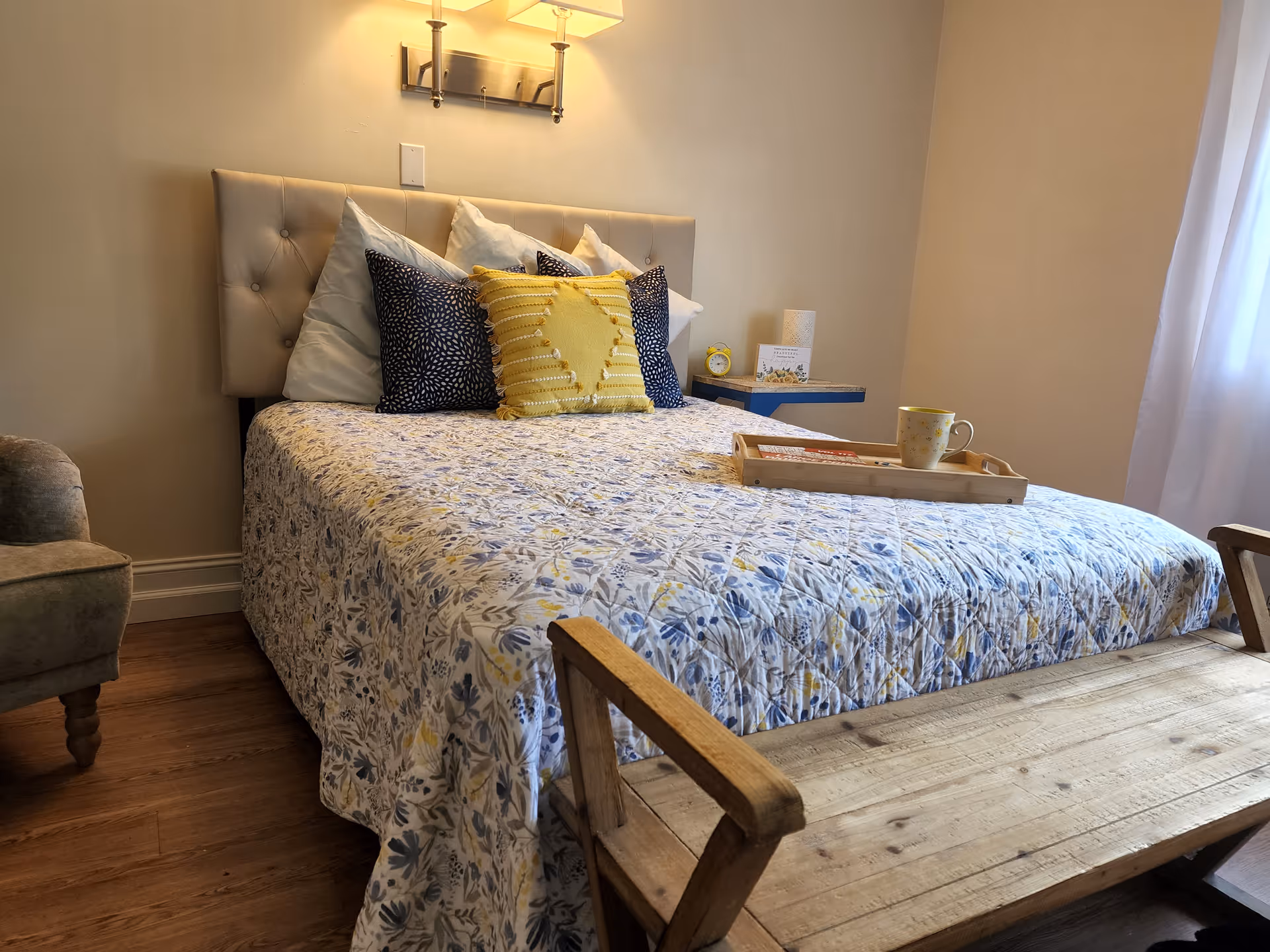 A cozy bedroom with a bed covered in a floral quilt and multiple pillows including a yellow decorative pillow. There is a wooden bench at the foot of the bed, a small side table with a yellow alarm clock and a card, and a wall-mounted light fixture above the headboard. Light filters in through a window with white curtains on the right side.