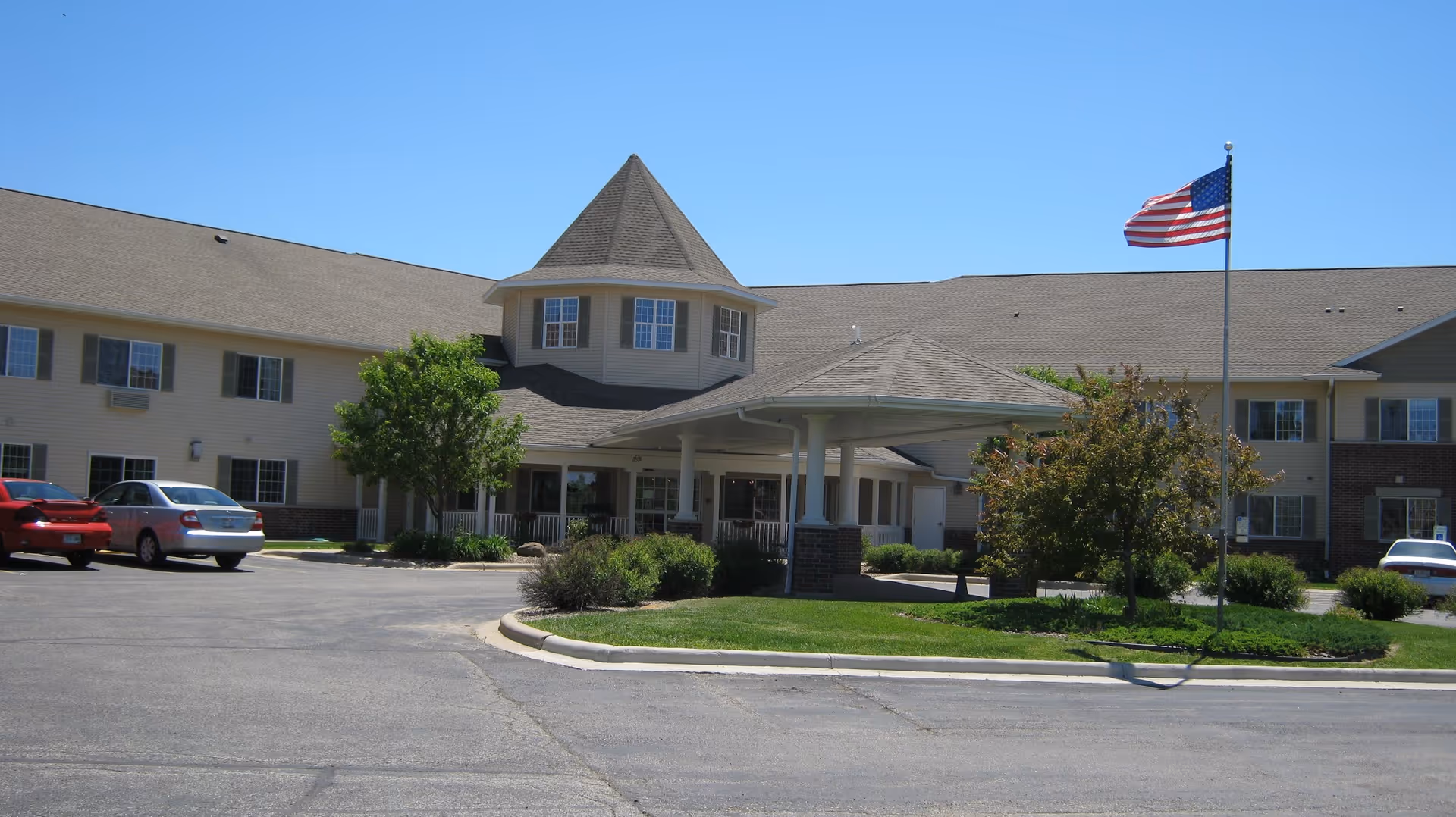 Exterior view of Drumlin Reserve - Assisted Living facility showing a large two-story building with a peaked roof and multiple windows. There is a covered entrance supported by columns, a flagpole with an American flag, some trees, shrubs, and a parking area with a few cars parked.