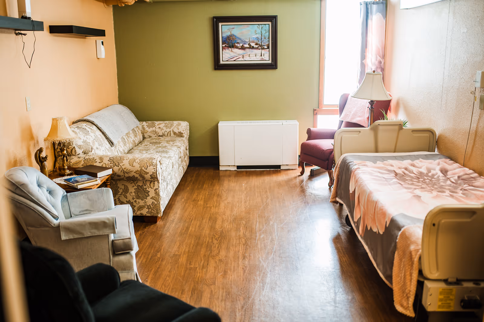 A cozy patient room in a senior care facility with bed, sofa, chair, and wood floor.