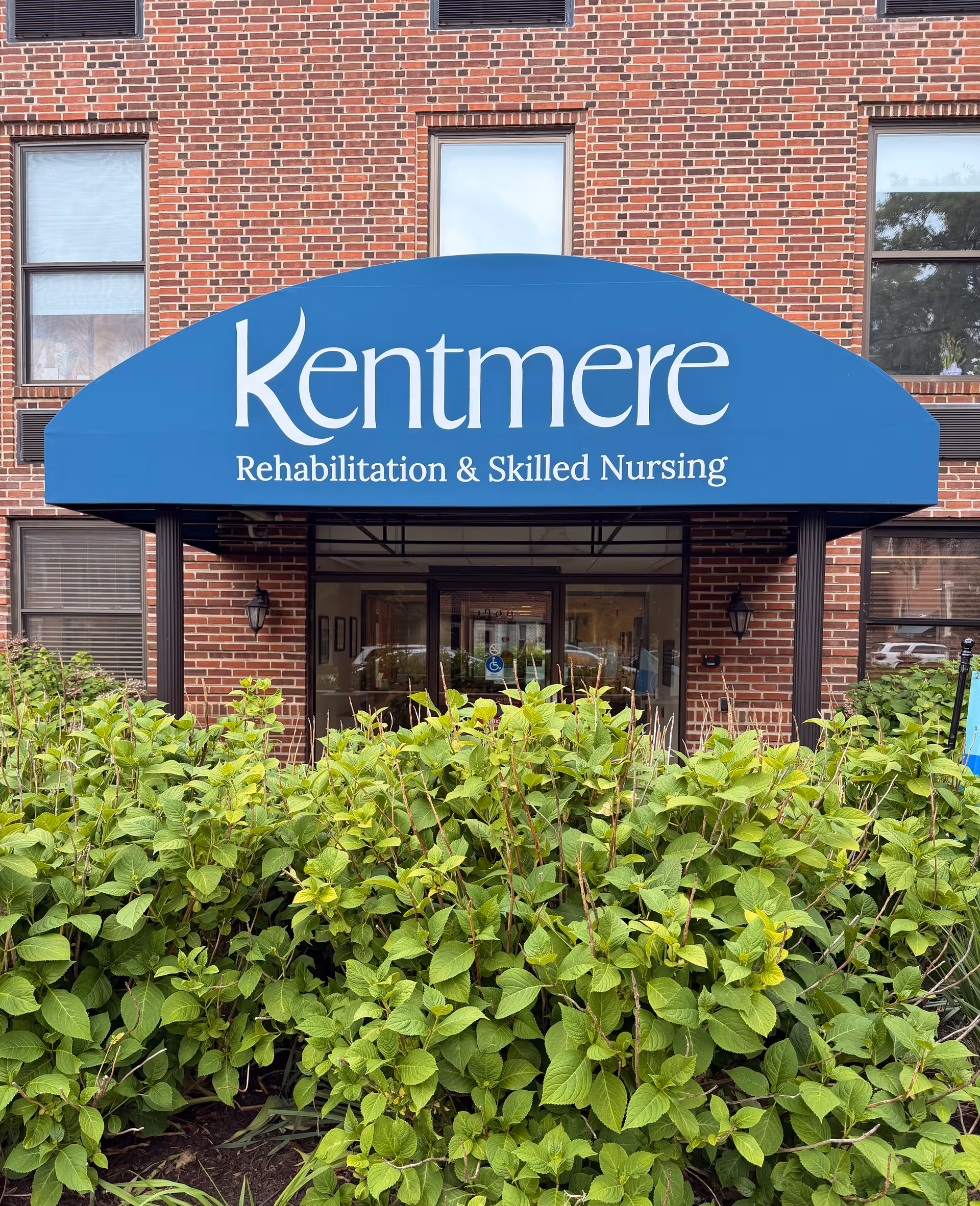 Brick building entrance with a blue Kentmere canopy sign and green shrubs.