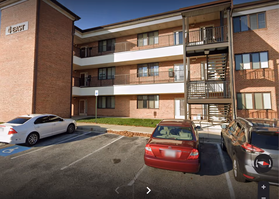 Exterior view of a brick senior living building with balconies and parked cars in front.