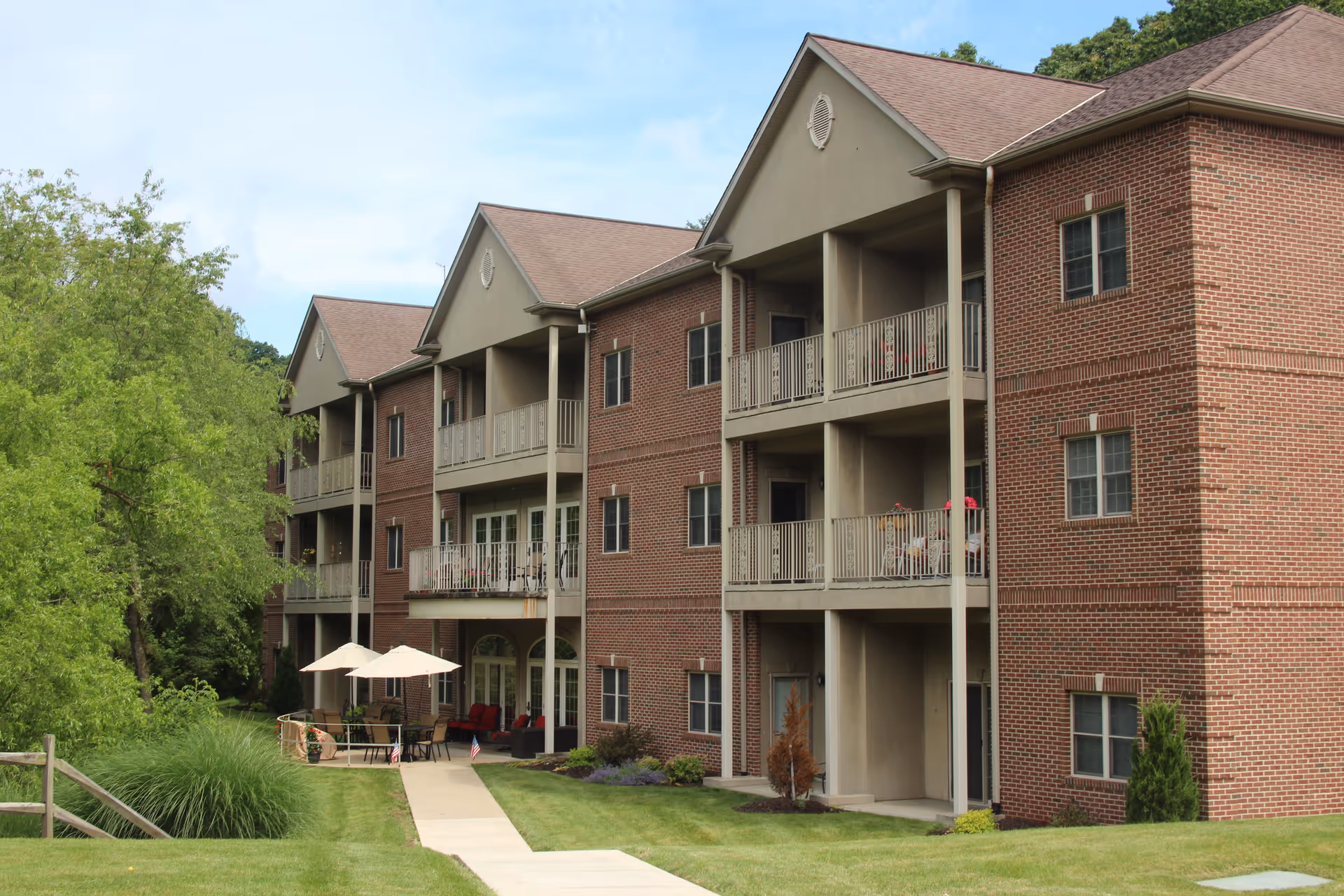 Three-story brick senior living building with balconies and a patio area with umbrellas set in landscaped grounds.