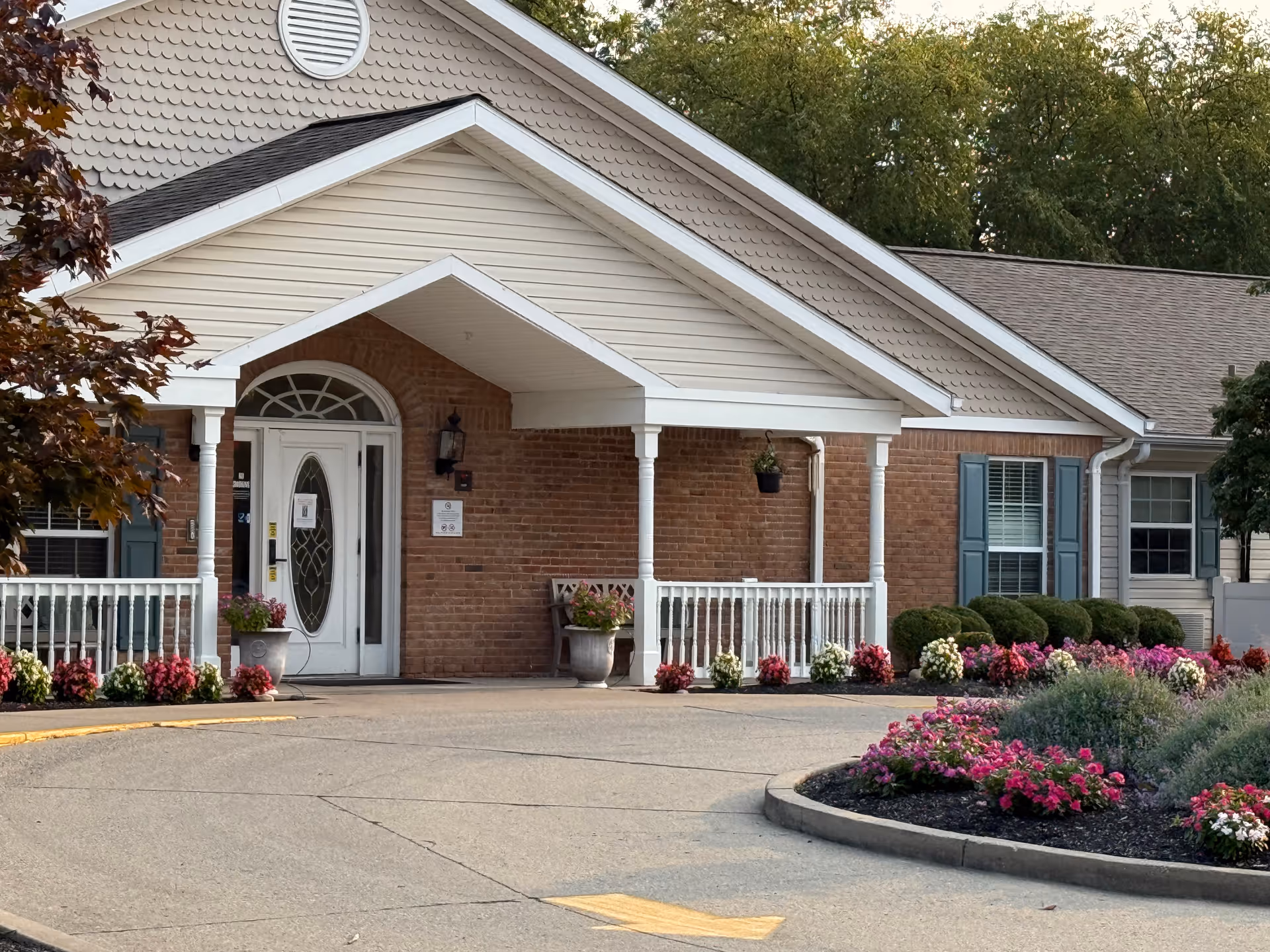 Brick senior living facility entrance with a covered porch and colorful flower beds.