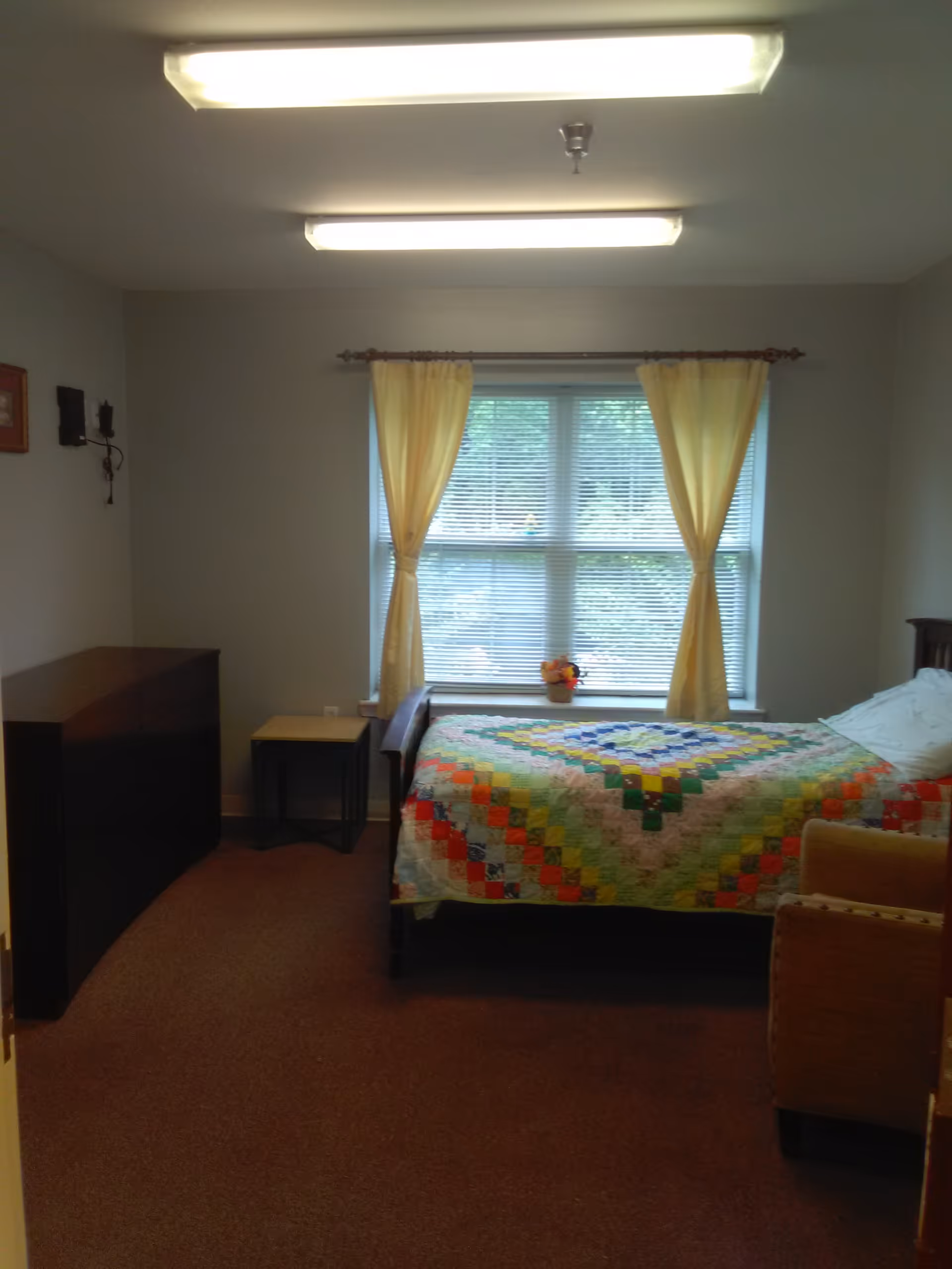A bedroom with a single bed covered by a colorful patchwork quilt, positioned against a wall with a window behind it. The window has yellow curtains tied back, and a small flower pot is placed on the windowsill. To the left of the bed, there is a small side table and a dark wooden dresser. The room has beige carpet and neutral-colored walls, with two fluorescent ceiling lights.