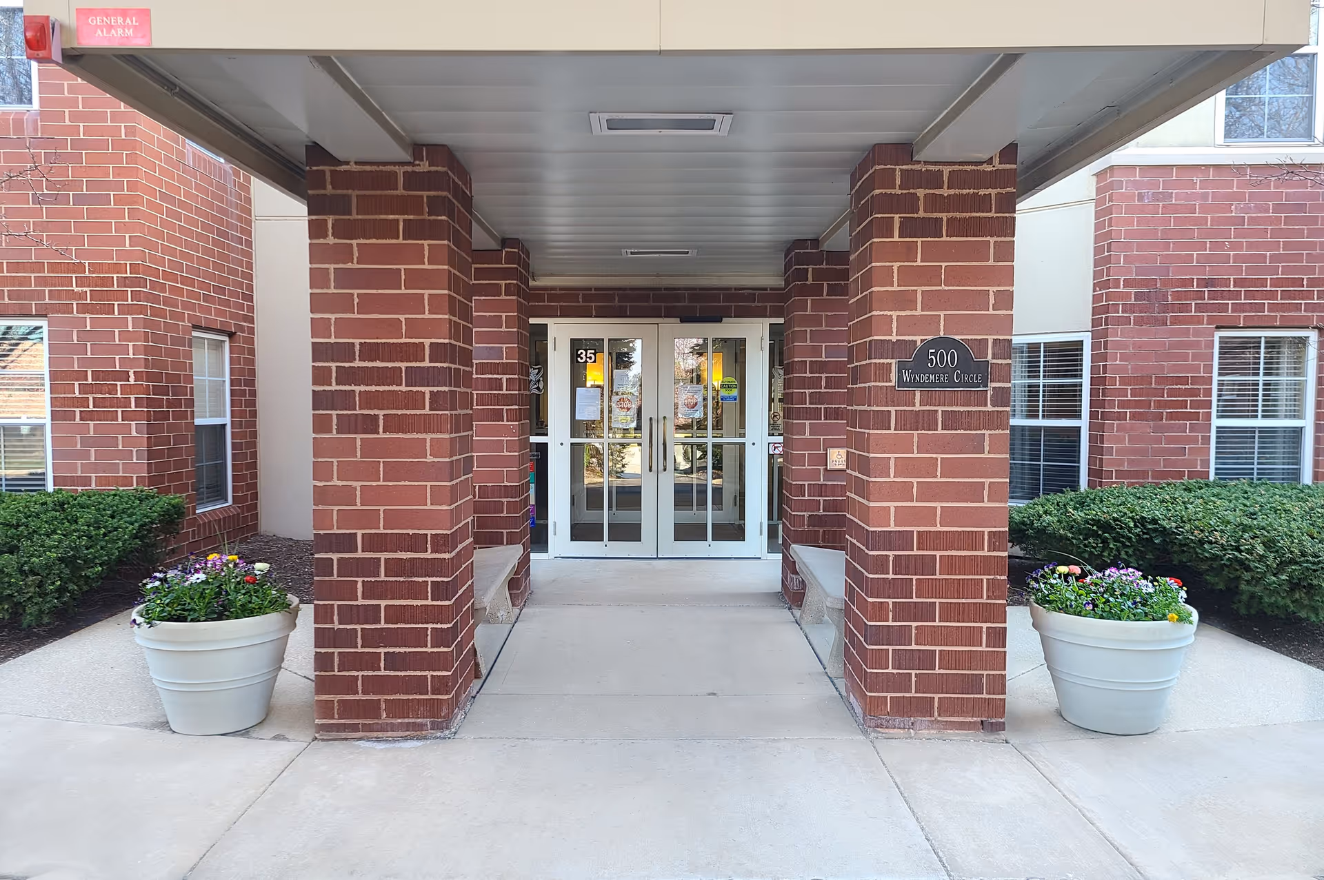 Entrance to a brick building with a covered walkway supported by brick columns. Two large white planters with colorful flowers are placed on either side of the walkway. Double glass doors are at the end of the walkway, with a sign on the right brick column that reads '500 Windemere Circle'.