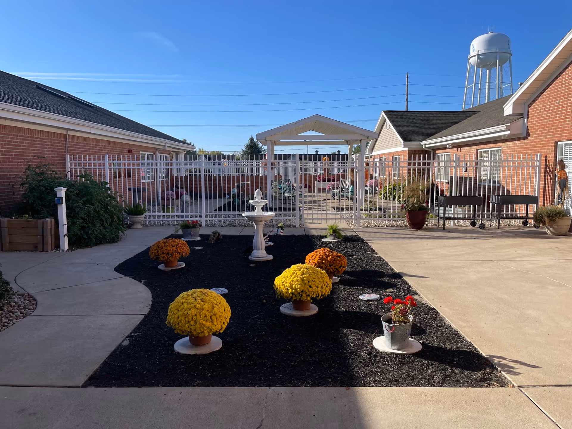Outdoor courtyard area at Woodbridge Health Campus featuring a small garden bed with yellow and orange potted flowers, a white decorative fountain, and a gated seating area with benches and chairs. The courtyard is surrounded by red brick buildings and a water tower is visible in the background under a clear blue sky.