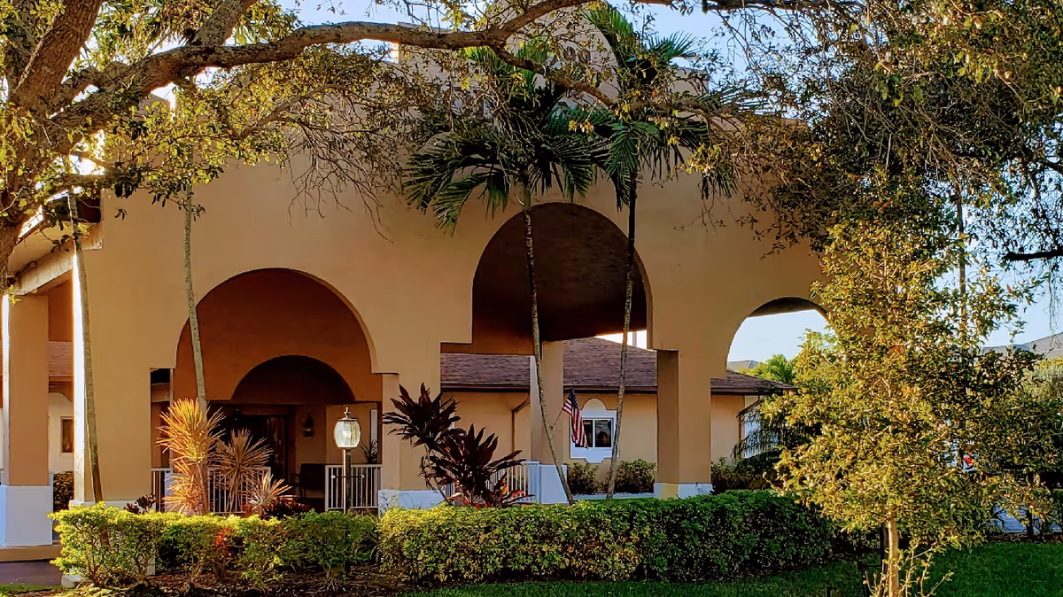 Front entrance of a beige building with large archways, palm trees, trimmed hedges and an American flag.