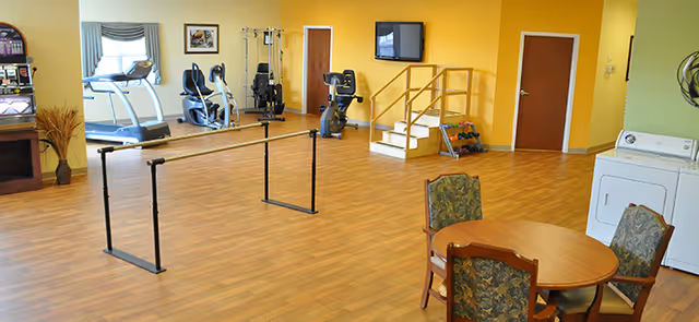 Open exercise room in a senior facility with treadmills, bikes, parallel bars, and a round table.