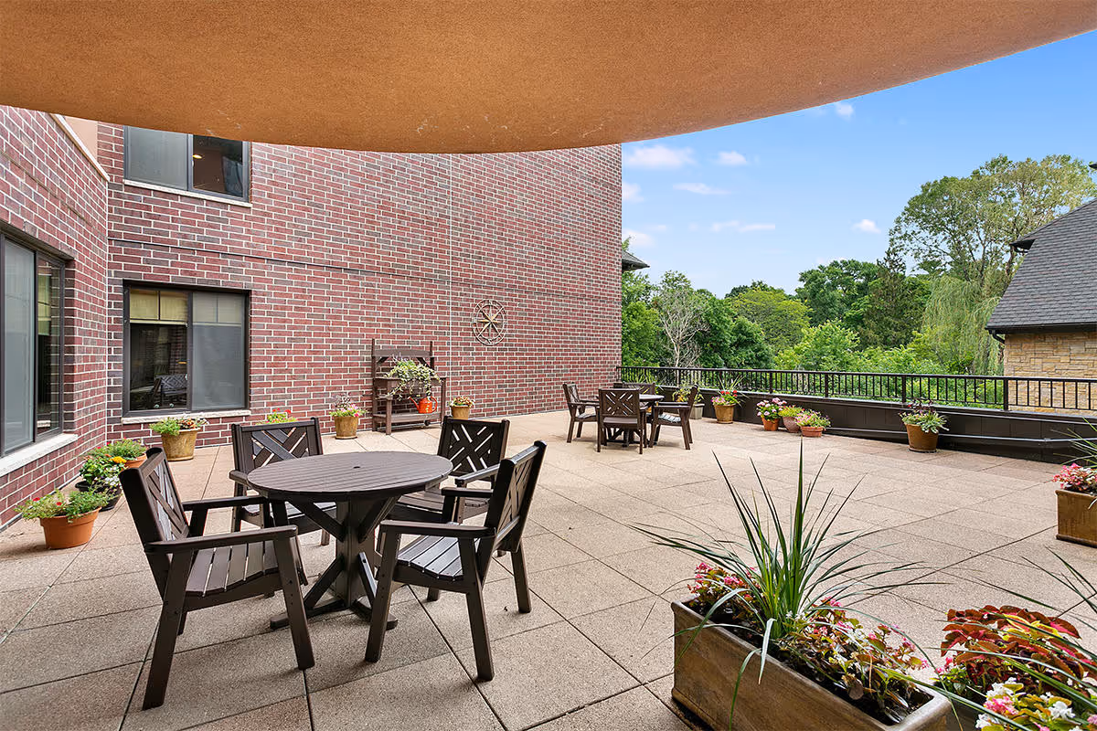 Outdoor patio area with multiple round tables and chairs, surrounded by potted plants and flowers. The patio is adjacent to a brick building and overlooks a green, tree-filled landscape under a blue sky with some clouds.