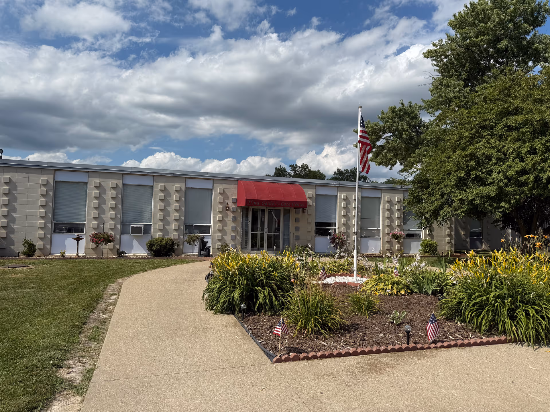 Low single-story senior living facility with a red awning over the entrance, a flagpole and landscaped flowerbeds in front under a partly cloudy sky.