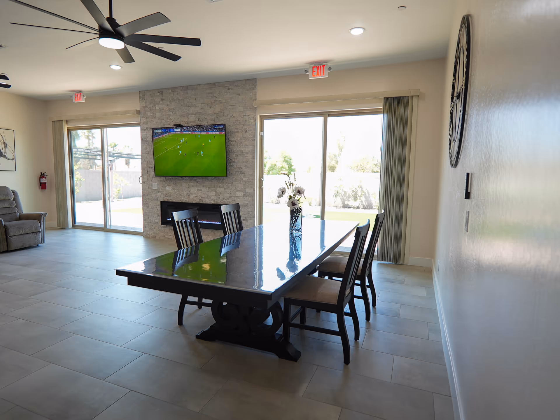 Bright common area with a long dining table, chairs, stone wall, and TV.