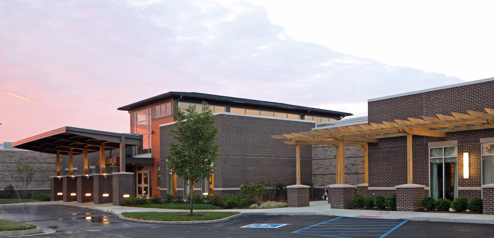 Front exterior of a modern brick senior living building featuring a covered entry, wooden pergola, landscaped island, and adjacent parking lot at sunset.