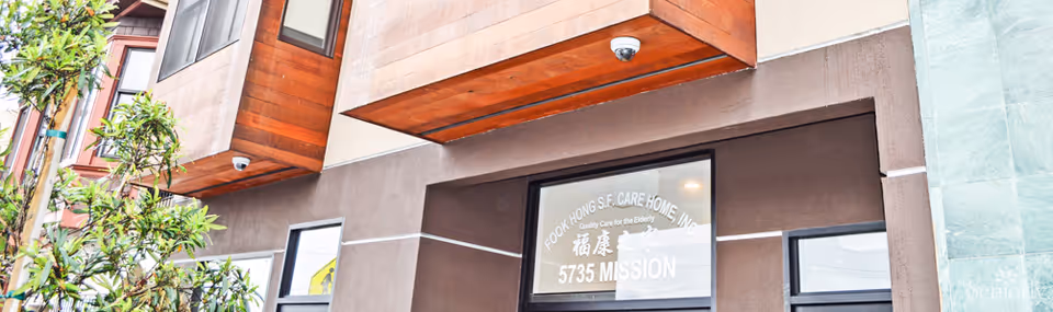 Exterior view of the entrance to Fook Hong Care Home, showing a modern building facade with brown and green walls, a security camera, and a sign on the door that reads 'Fook Hong S.F. Care Home Inc, Quality Care for the Elderly, 5735 Mission'.