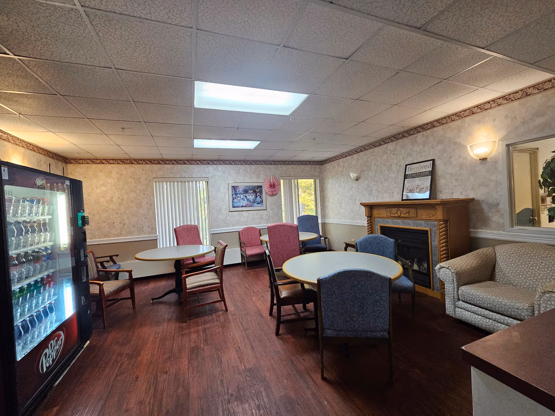 Interior senior living lounge with round tables, chairs, a fireplace, sofa, and vending machine.