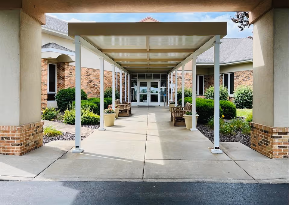 Covered outdoor walkway with white columns, benches, and planters leading to the brick facility entrance.