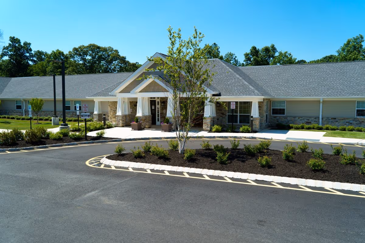 Front entrance of a single-story senior living building with a gabled portico, landscaped circular driveway, and small trees and shrubs.