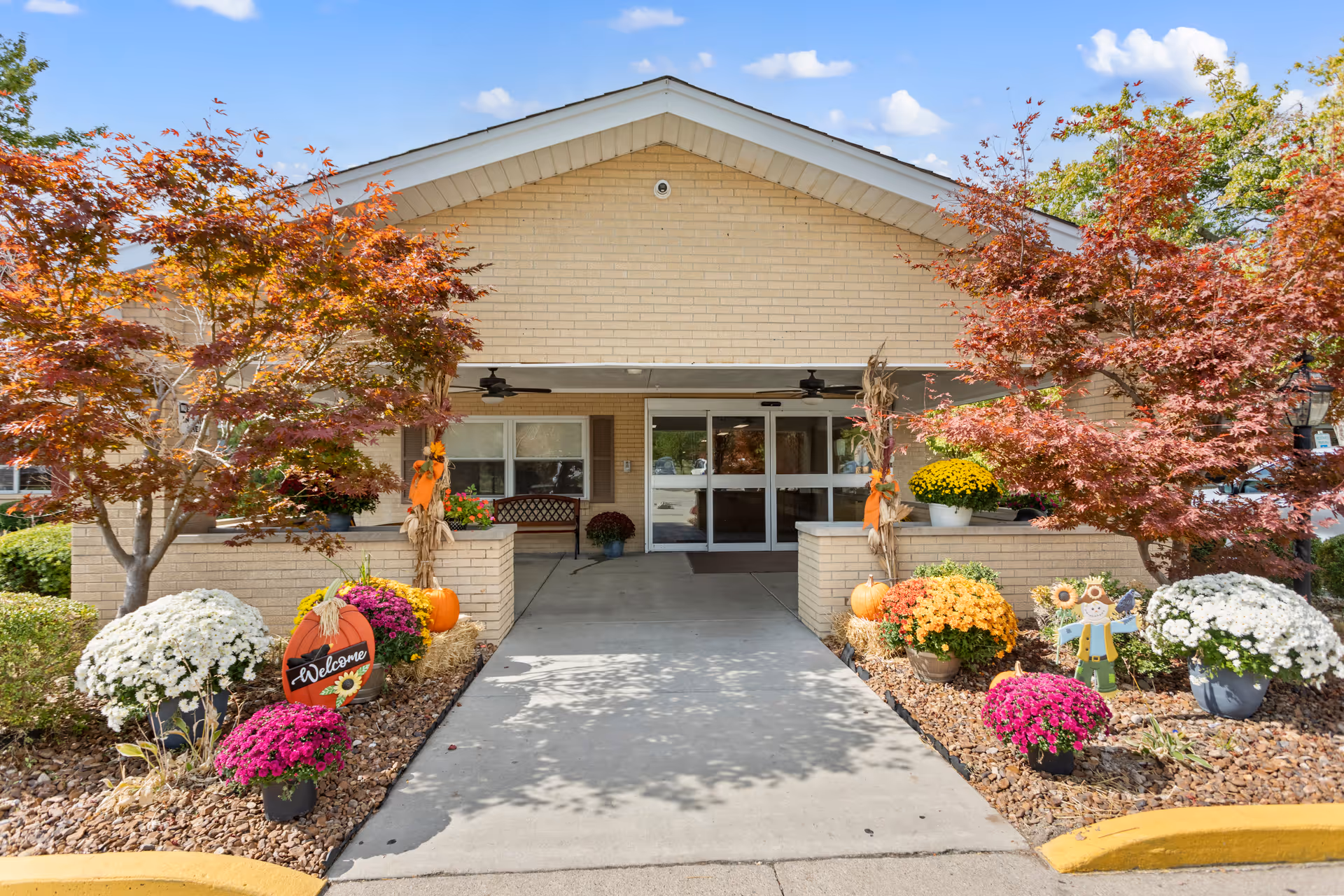 Entrance to a nursing care center with a covered walkway, decorated with autumn-themed plants, pumpkins, and a welcome sign. Trees with red and orange leaves flank the walkway leading to glass double doors.