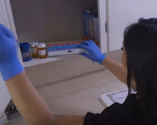 Caregiver wearing blue gloves preparing medications from a cabinet on a countertop.