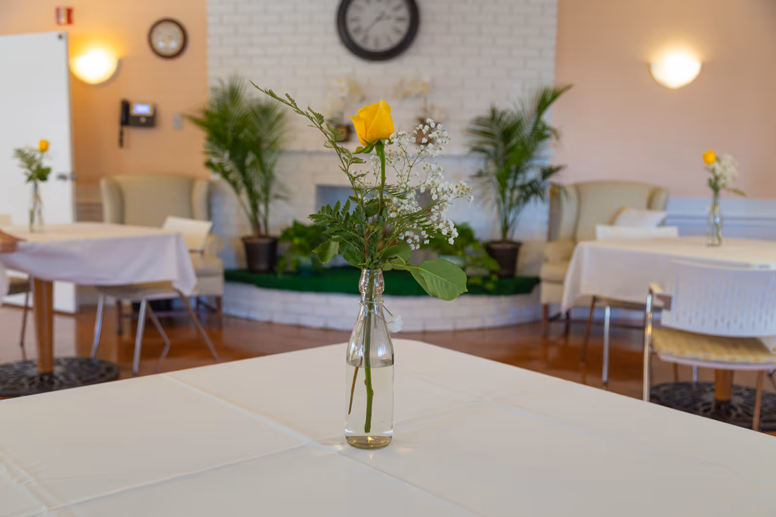 A dining area with tables covered in white tablecloths, each table featuring a small glass vase with a yellow rose and white baby's breath flowers. In the background, there are beige armchairs, potted plants, a white brick fireplace, and a wall clock.
