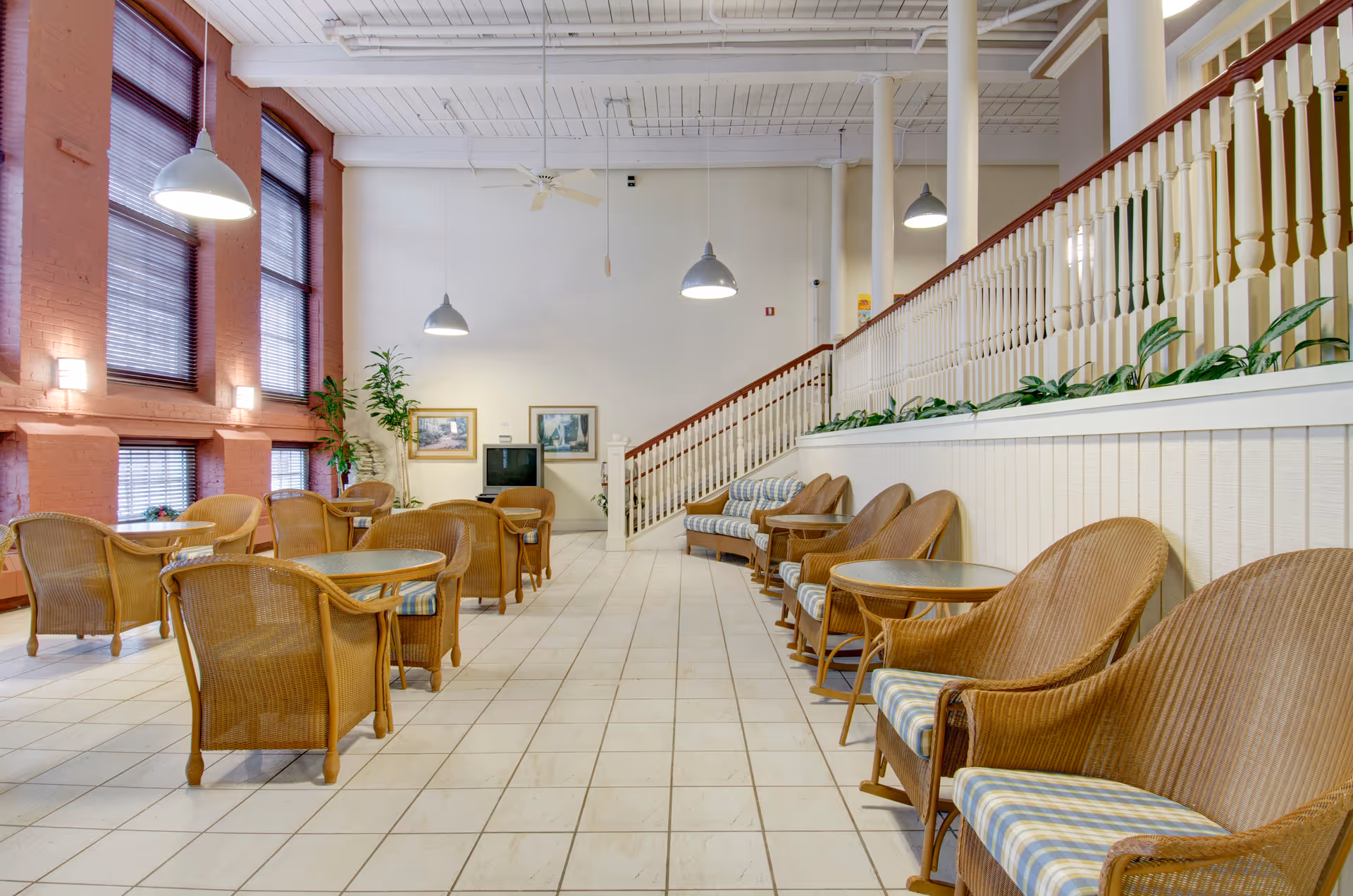Bright communal lounge with wicker chairs and small tables, high ceilings, large windows, and a staircase with railing along the right side.