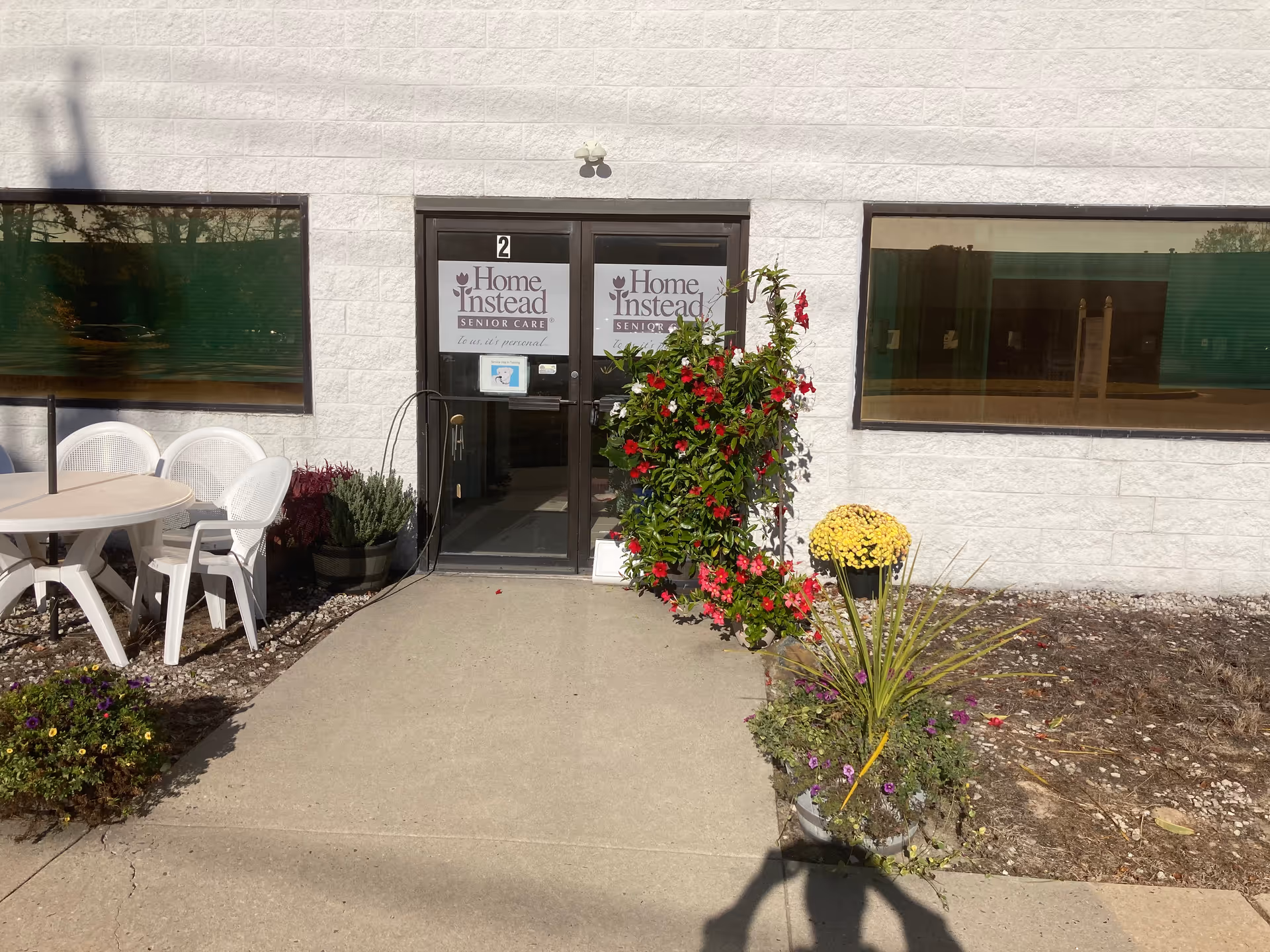Exterior facility entrance with double doors, Home Instead Senior Care sign, and outdoor seating.